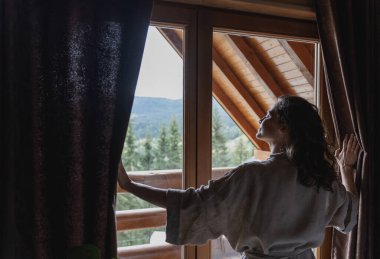 Dağlardaki ahşap dağ evinde perdeleri açan beyaz bir kadın. Doğadaki bir kütük evde rahat bir dinlenme.