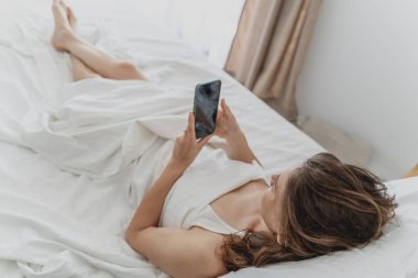 Sabahları beyaz çarşafların üzerinde uzanmış, cep telefonunun ekranına bakan genç bir kadın.