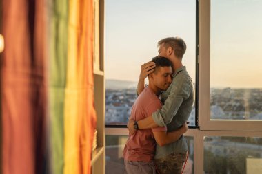 Pencerenin yanında şehre bakan bir dairede kucaklaşan genç, sevimli eşcinsel çift. LGBT Ailesi