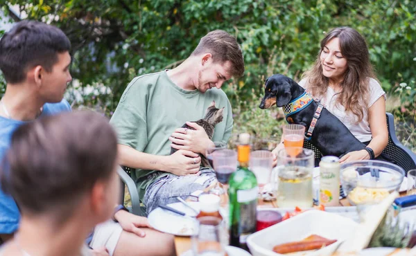 A group of young people friends sitting at a table at a summer garden party with pets kitten and dachshund dog on their laps