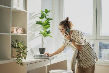 Genç, beyaz, gözlüklü, dizüstü bilgisayarda çalışan ve pencerenin yanındaki modern bir apartman dairesinde akıllı telefon kullanan bir kadın.