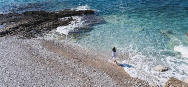Mavi okyanus kıyısında dalgalarla yürüyen beyaz gömlekli genç bir kadın. Tatiller ve tatiller