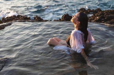 Deniz kıyısında doğal bir deniz banyosunda beyaz gömlekli, neşeli, rahat bir kadın. Thalasapi sağlıklılığı