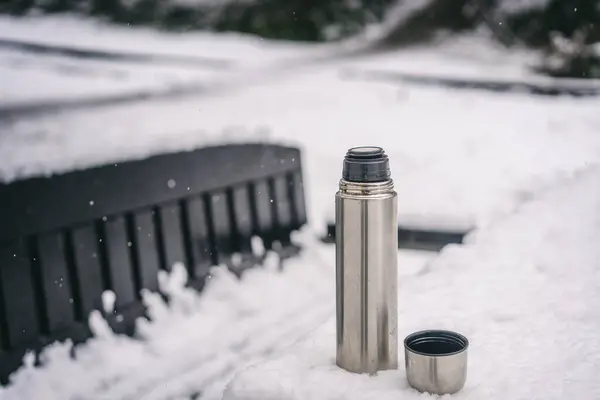 A thermos with a warm drink on a table in a winter park in snowy weather. Healthy lifestyle concept