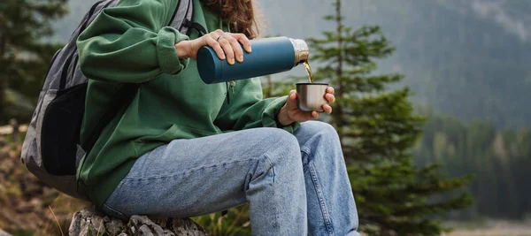 Woman traveler pours tea from a thermos while sitting in the forest