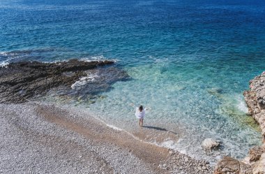 Mavi okyanus kıyısında dalgalarla yürüyen beyaz gömlekli genç bir kadın. Tatiller ve tatiller