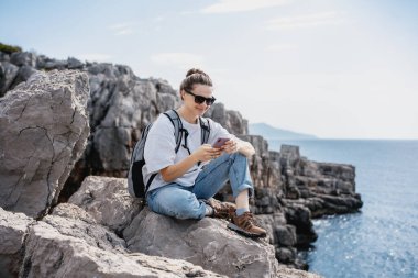 Gezgin bir kadın elinde akıllı bir telefonla kayalık bir deniz kıyısında dinleniyor. Yaz seyahati ve macera