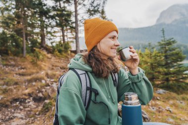 Sırt çantası ve termosuyla dağlardaki gölün kıyısında gezen neşeli genç bir kadın.