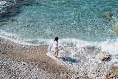 Mavi okyanus kıyısında dalgalarla yürüyen beyaz gömlekli genç bir kadın. Tatiller ve tatiller