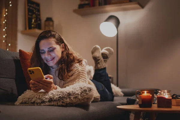 A young woman spends a cozy winter evening at home lying on the sofa looking at her smartphone. Winter holidays, Christmas and online surfing
