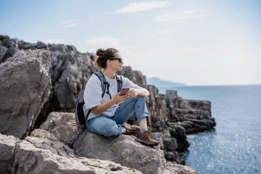 Gezgin bir kadın elinde akıllı bir telefonla kayalık bir deniz kıyısında dinleniyor. Yaz seyahati ve macera