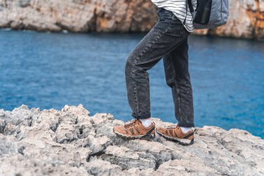 Kayalık bir sahil boyunca yürüyüş botlarıyla yürüyen bir kadın gezgin. Yürüyüş ve macera konsepti