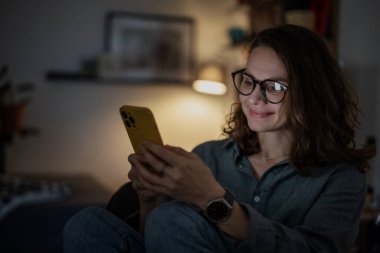 Genç bir kadın akşamları evde otururken akıllı telefon ekranına bakıyor.