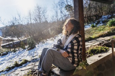 Bir kır evinin terasında sıcak bir battaniyeyle güneşin tadını çıkaran genç bir kadın.