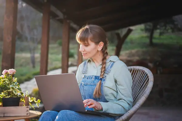 春天的一天 身穿粗斜纹棉布工作服的年轻而快乐的女人坐在乡村房屋的阳台上 手里拿着笔记本电脑 图库照片