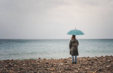 Genç bir kadın deniz kıyısında mavi bir şemsiyeyle yağmurda yürüyor.
