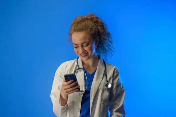 青い背景の携帯電話を使って若い女性医師のスタジオショット — ストック写真