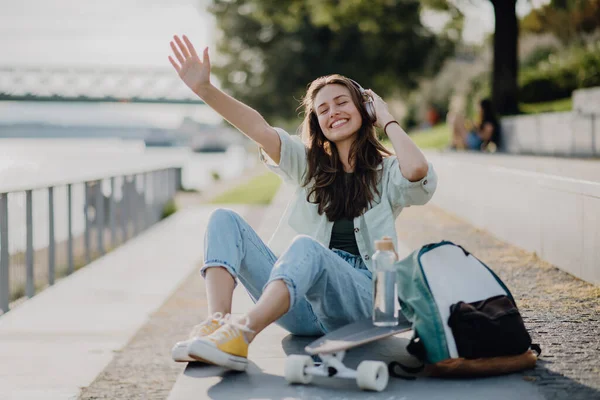 スケートボードに座って街の橋で音楽を聴いている幸せな若い女性 若者文化と通勤概念 — ストック写真