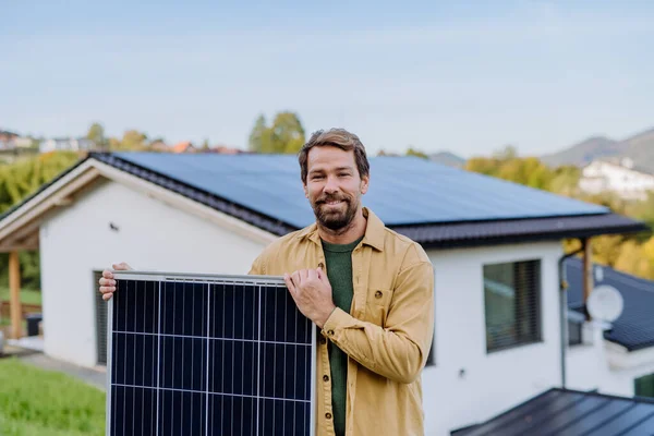 Mature man holding a solar panel near his house with solar panels on the roof. Alternative energy, saving resources and sustainable lifestyle concept.