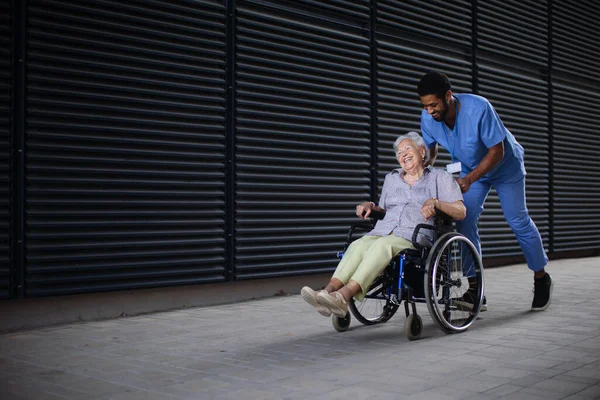 路上の車椅子で興奮したシニア女性を押して一緒に時間を楽しむ介護者の男 — ストック写真
