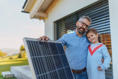 Babası ve küçük kızı güneş paneli olan evin yakınlarında. Alternatif enerji, tasarruf kaynakları ve sürdürülebilir yaşam tarzı kavramı.