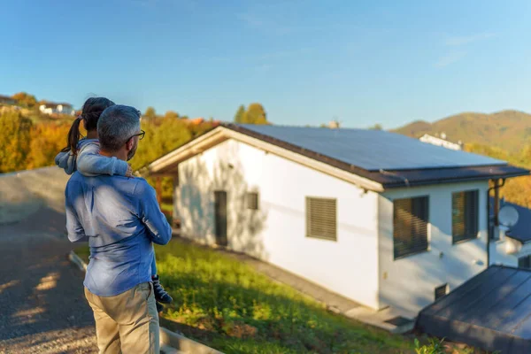 回头看爸爸抱着她的小女孩 用太阳能板看着她们的房子 替代能源 节约资源和可持续的生活方式概念 — 图库照片