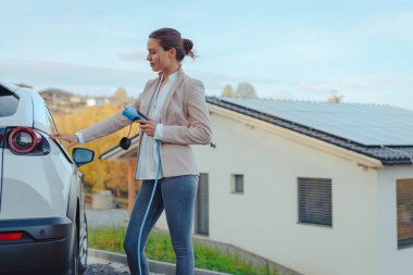 Evde elektrikli araba şarj eden genç bir kadın, sürdürülebilir ve ekonomik ulaşım kavramı.