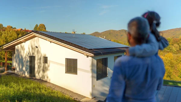 お父さんは腕の中で彼女の小さな女の子を保持し 太陽電池パネルで彼らの家を見ての背面ビュー — ストック写真