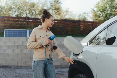 Arabasından elektrik kablosu tutan genç bir kadın, onu evde şarj etmeye hazır, sürdürülebilir ve ekonomik ulaşım konsepti.