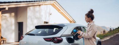 Arabasından elektrik kablosu tutan genç bir kadın, onu evde şarj etmeye hazır, sürdürülebilir ve ekonomik ulaşım konsepti.