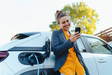 Akıllı telefonlu genç bir kadın elektrikli araba şarj istasyonunda beklerken, sürdürülebilir ve ekonomik ulaşım kavramı.