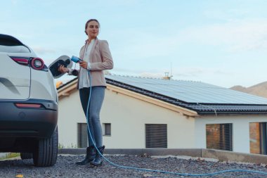 Evde elektrikli araba şarj eden genç bir kadın, sürdürülebilir ve ekonomik ulaşım kavramı.