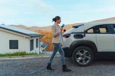 Arabasından elektrik kablosu tutan genç bir kadın, onu evde şarj etmeye hazır, sürdürülebilir ve ekonomik ulaşım konsepti.