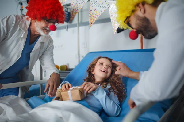 Happy doctors with clown red noses celebrating birthday with little girl in a hospital room.