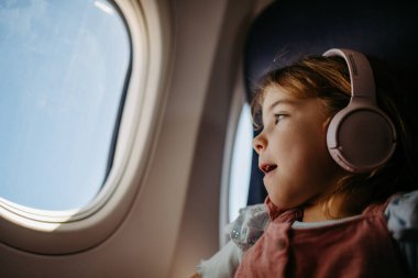 Little girl in airplane listening music and looking out of the window. clipart