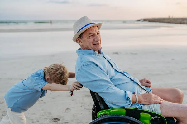 車椅子で祖父を押して一緒に海を楽しむ男の子 — ストック写真