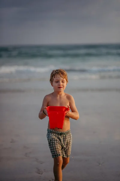 Little boy in swimsuit with bucket full of water running out of sea, enjoying summer holiday.