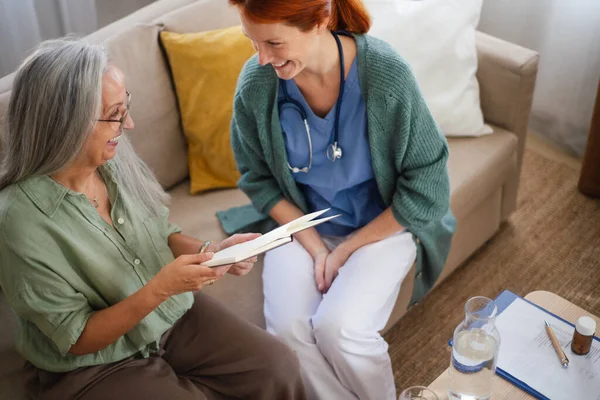 Senior woman and nurse reading book together, having nice time at home.