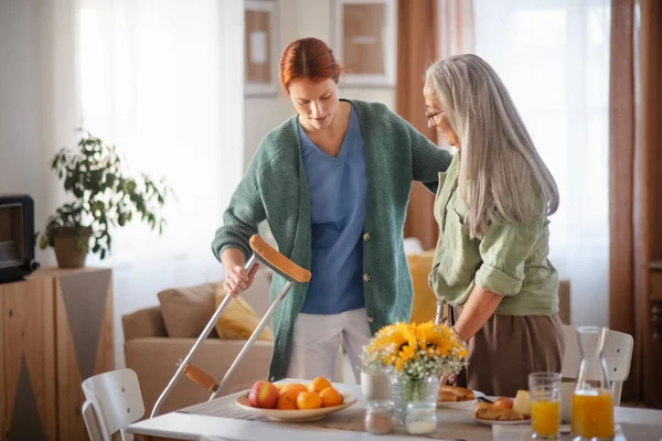 Nurse helping senior woman with walking after leg injury, in her home.