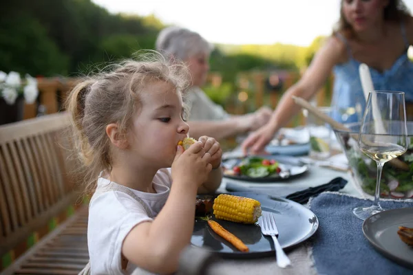 A family eating at barbecue party dinner on patio, little girl eating roasted corn and enjoying it.