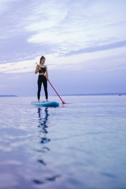 A young beautiful girl surfer paddling on surfboard on the lake at sunrise