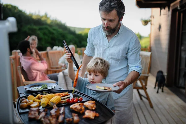 夏の庭のパーティー中に小さな息子のグリルリブと野菜のグリルを持つ父親 — ストック写真