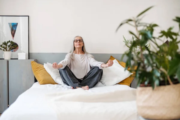 stock image Senior woman doing yoga and meditating on a bed.