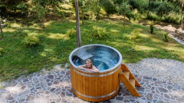 Young woman enjoying wooden bathtub with fireplace to burn wood and heat water in backyard in mountains. High angle view.
