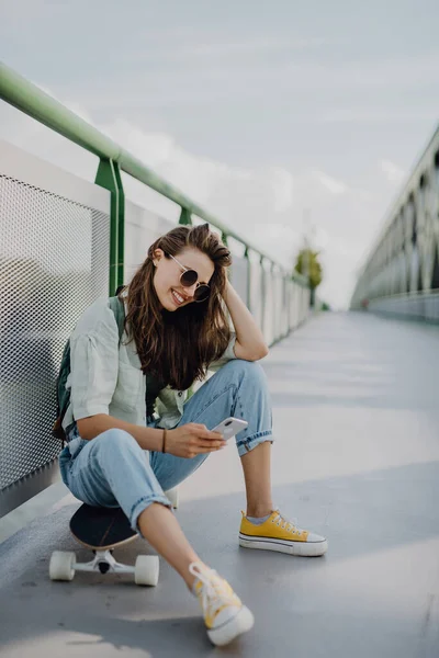 スケートボードに座って街の橋でスマートフォンをスクロールする幸せな若い女性 若者文化と通勤概念 — ストック写真