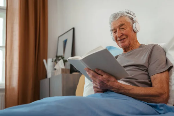 Старший Чоловік Розслабляється Ліжку Вивчає Книгу Слухає Музику Через Навушники — стокове фото