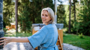A happy young woman standing in garden near hot tub and enjoying cup of morning coffee on summer vacation in mountains.