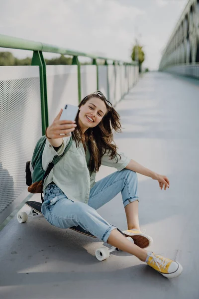 スケートボードに座って街の橋で自撮りした幸せな若い女性 若者文化と通勤概念 — ストック写真