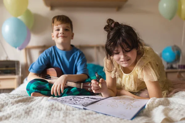 小男孩在吉他上敲打 而他的姐姐在床上画了一幅画 — 图库照片