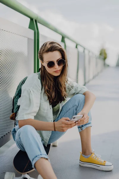 スケートボードに座って街の橋で携帯電話をスクロールする幸せな若い女性 若者文化と通勤概念 — ストック写真